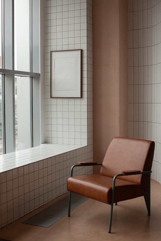 Blank frame hanging on tiled wall near window in small light room with soft brown leather armchair on floor at home